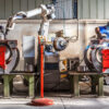 The mechanical performs welds metal components assisted by a worker.
